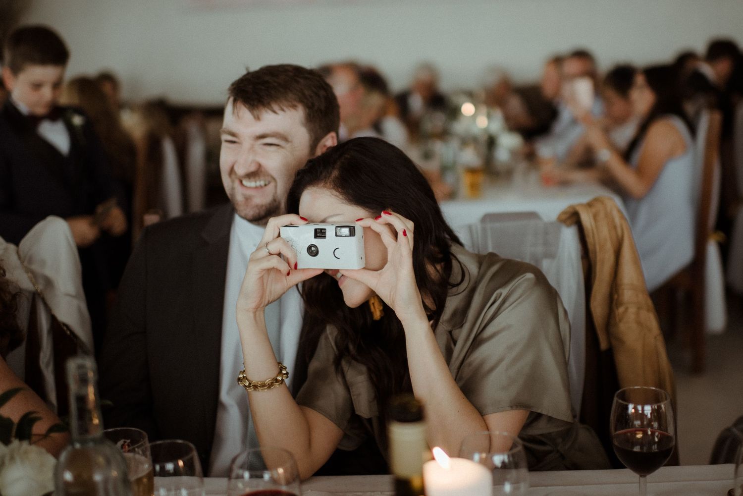 Wedding guests share a candid moment at a reception as someone takes a photo with a vintage point-and-shoot camera at Villa Malla in Filtvet