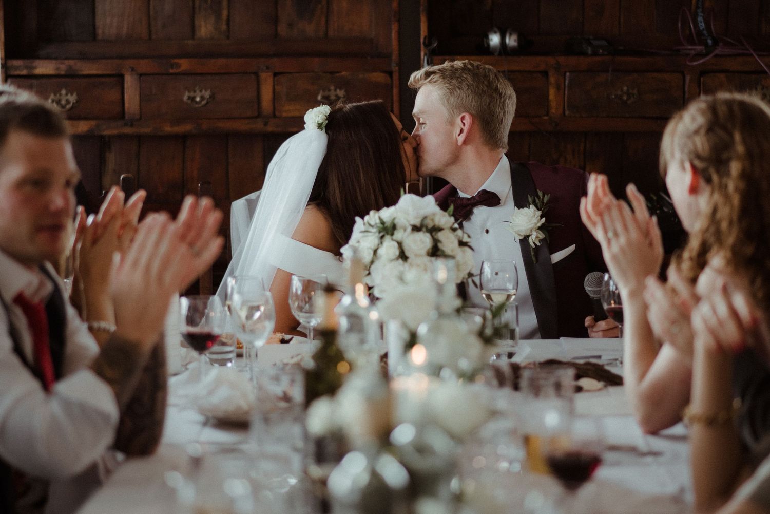 Wedding couple kisses at reception while guests clap at long wooden table decorated with flowers at Villa Malla in Filtvet, Norway.