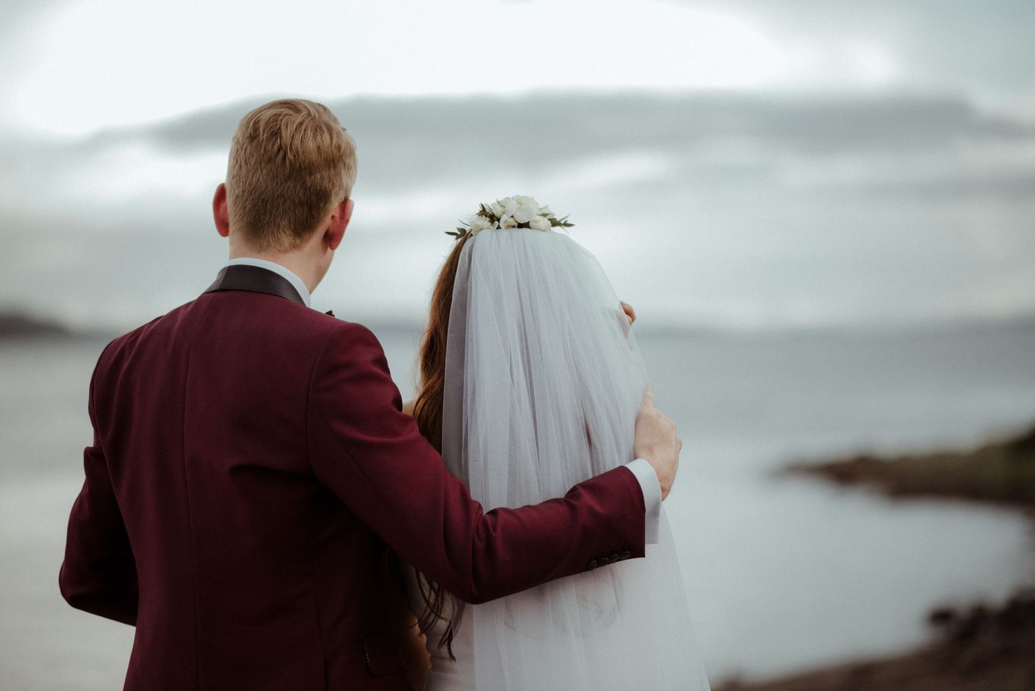 A couple in wedding attire embrace while looking out over a misty coastal landscape in muted tones at Villa Malla in Filtvet, Norway.