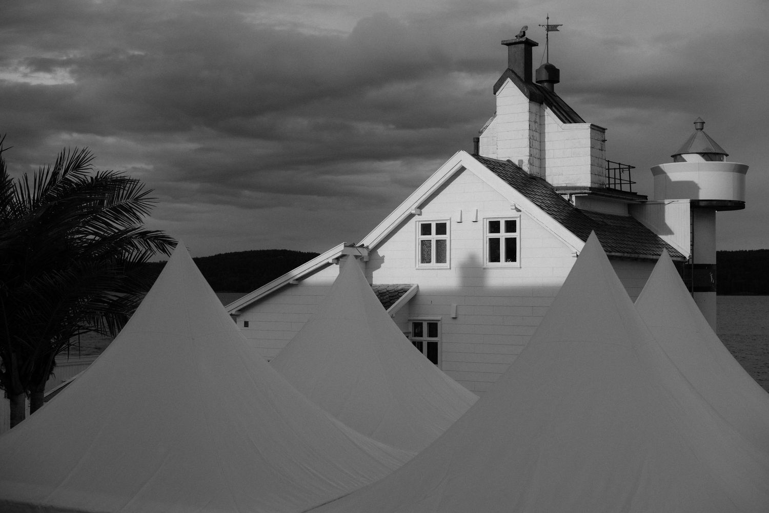 Black and white architectural photo showing white peaks of tents against a house with a dramatic cloudy sky at Villa Malla in Filtvet.