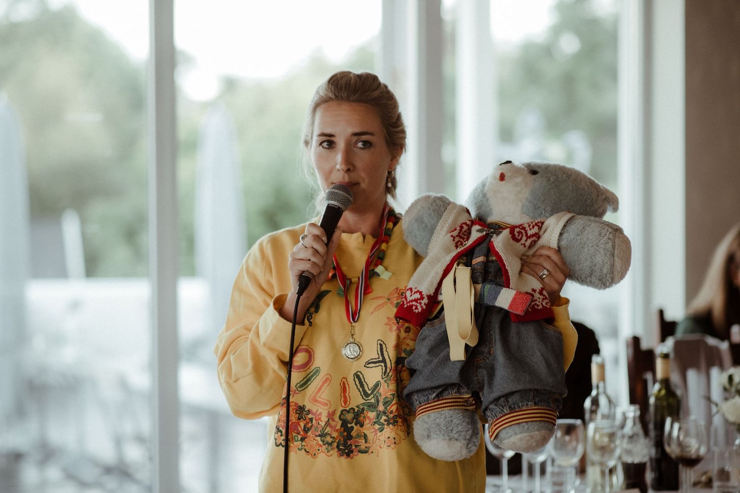 Someone in yellow floral attire holds a beverage and stuffed animal toy while standing by bright windows at Villa Malla in Filtvet, Norway.