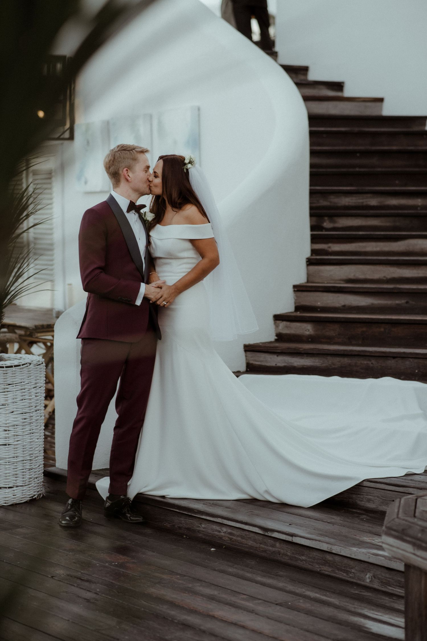 A couple shares an intimate moment on wooden stairs, with the bride's white off-shoulder gown trailing behind at Villa Malla in Filtvet.
