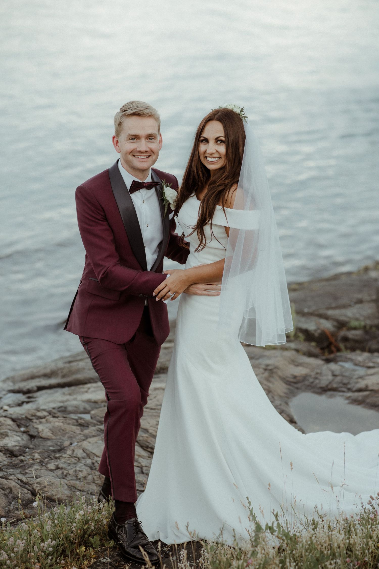 A wedding couple poses on rocky coastal cliffs with the groom in burgundy suit and bride in flowing white dress at Villa Malla in Filtvet.