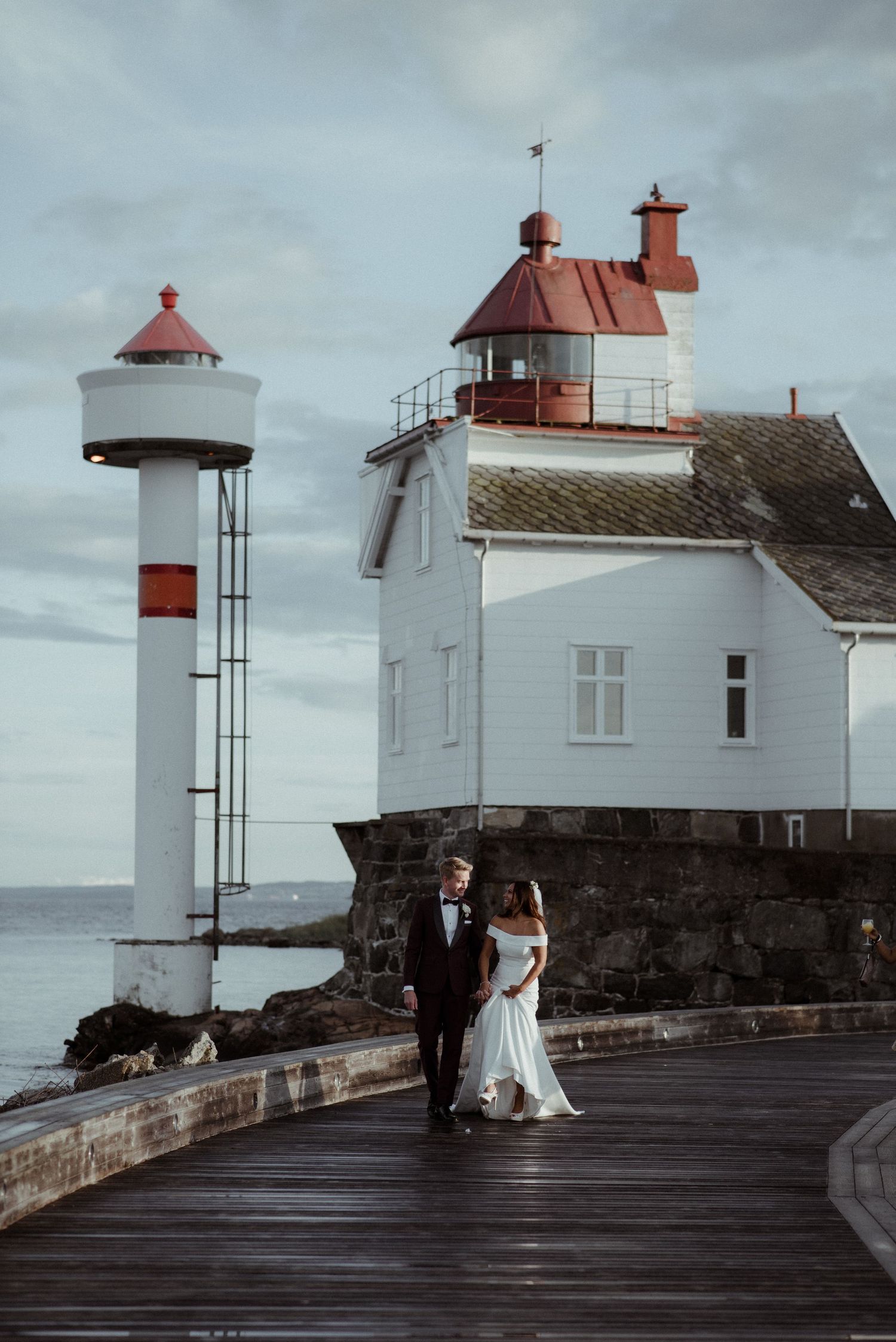 A wedding couple poses by a white lighthouse with red accents against a moody sky at Villa Malla in Filtvet, Norway.