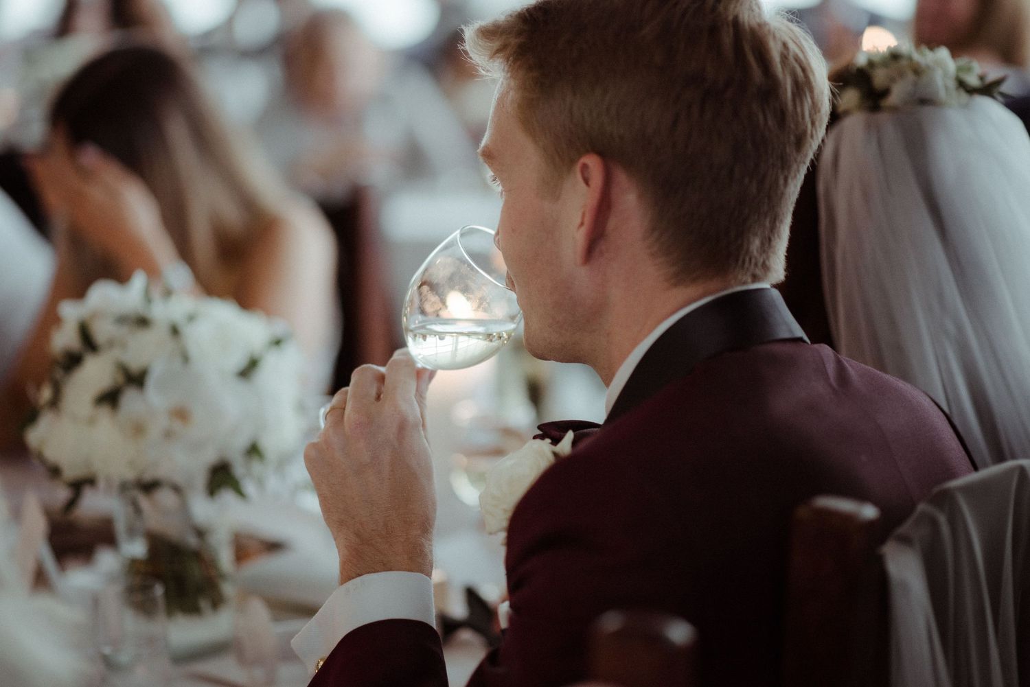The groom in burgundy suit sips from a wine glass at an elegant wedding reception at Villa Malla in Filtvet, Norway.