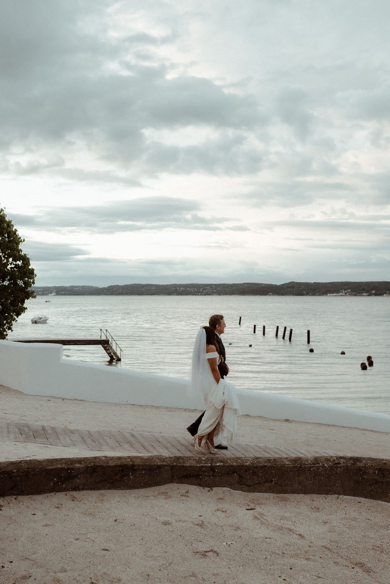 Newlyweds walks along a misty shoreline with old wooden posts in the water under cloudy skies at Villa Malla in Filtvet, Norway.