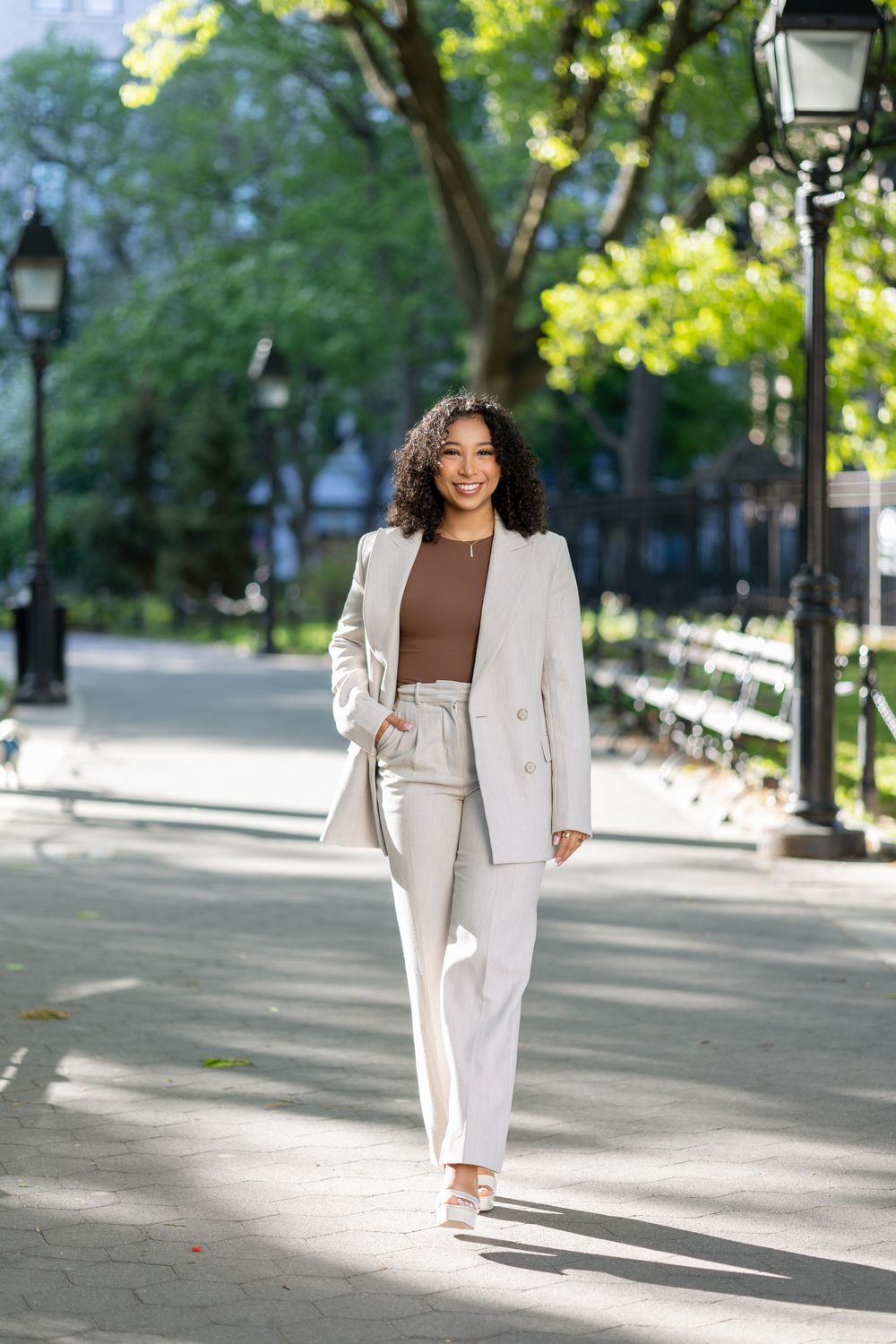 Woman in cream pantsuit walks confidently through park pathway on sunny day.