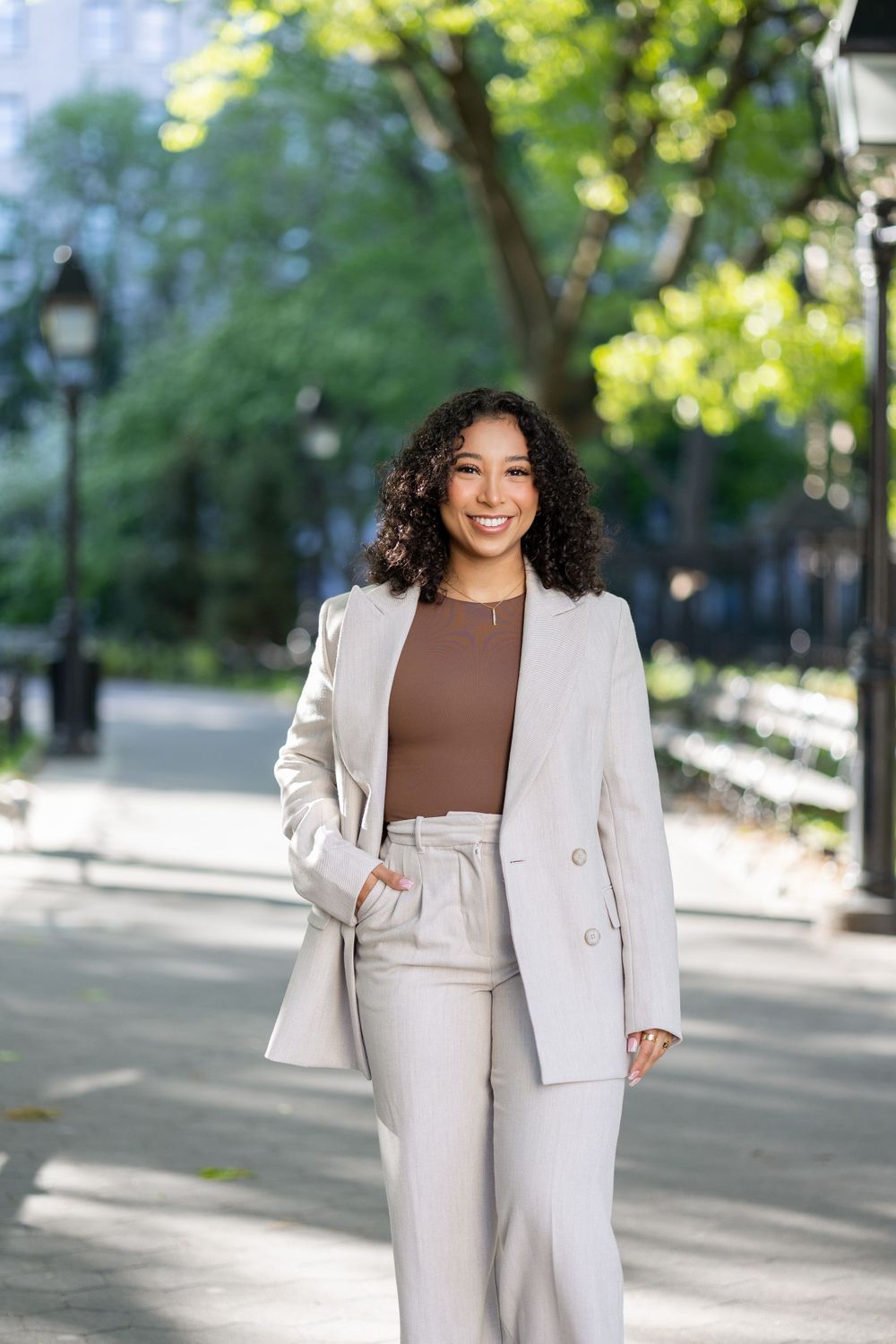 Woman in beige business suit poses casually on tree-lined pathway.