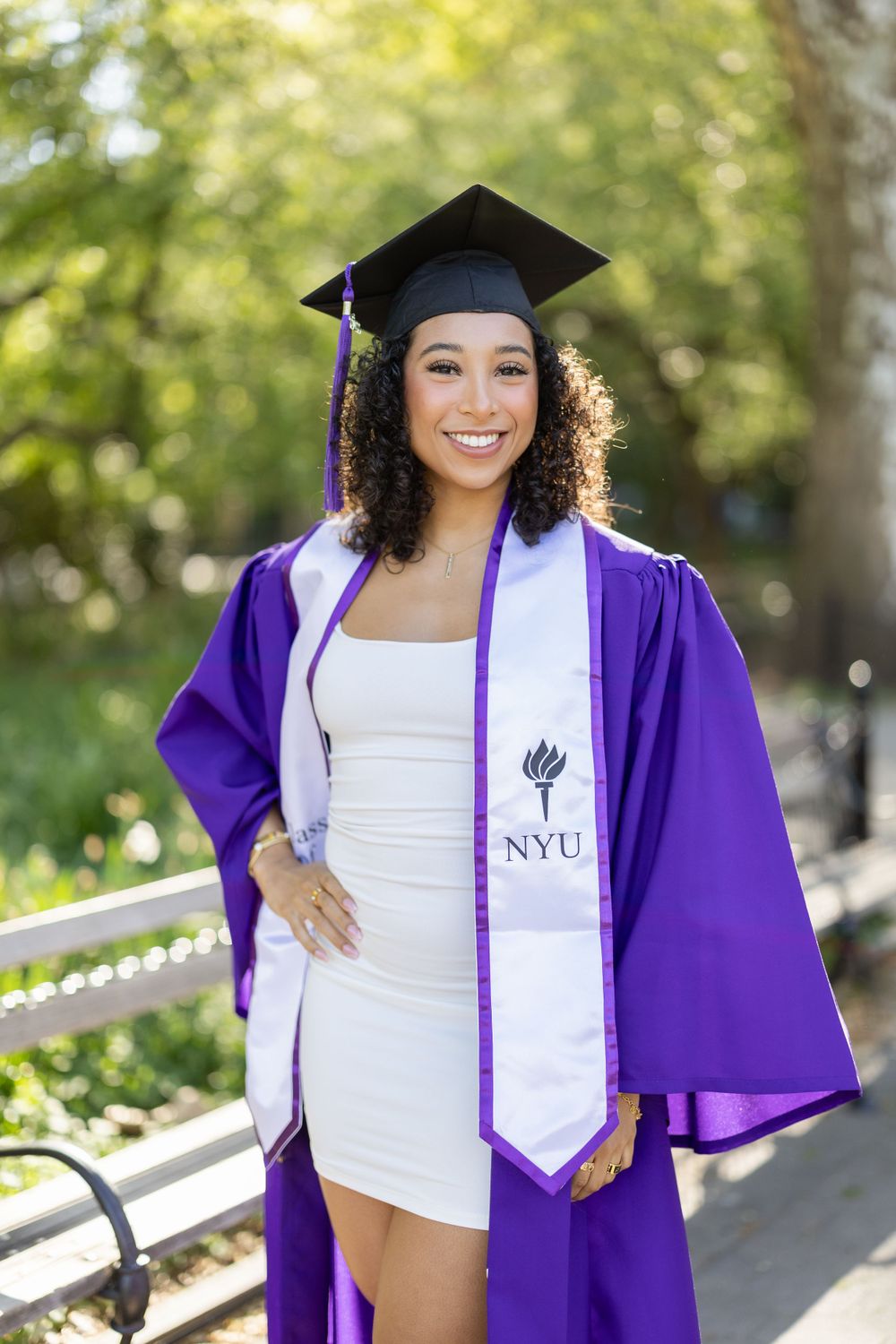 Graduate wearing purple NYU graduation gown and cap poses outdoors with natural bokeh background.