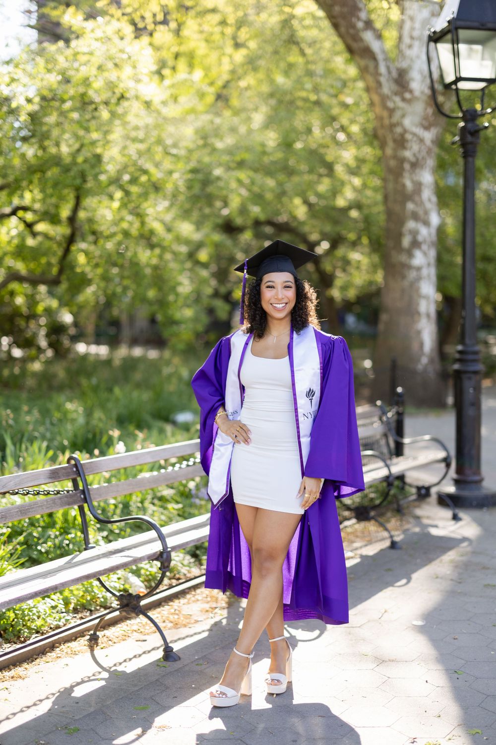 Graduate in purple academic regalia poses on park bench during sunny graduation photoshoot.