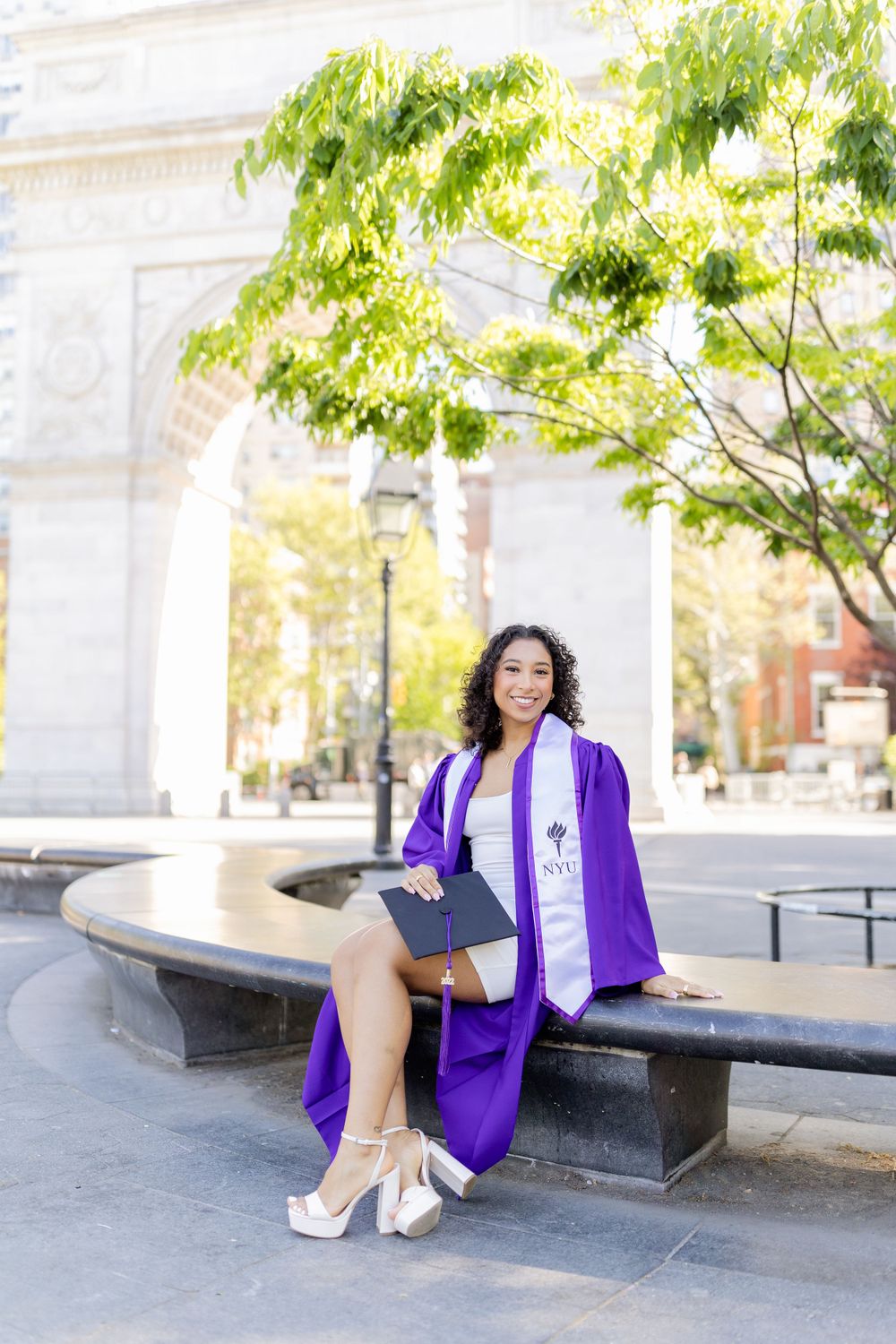 Graduate in purple regalia sits on bench near classical architecture on campus.