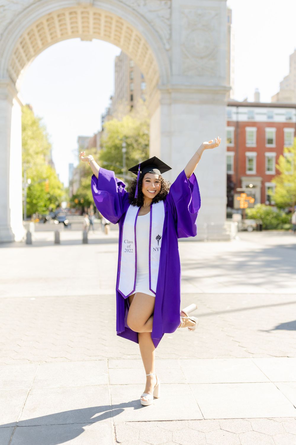 Graduate jumps joyfully in purple graduation gown near historic stone archway.