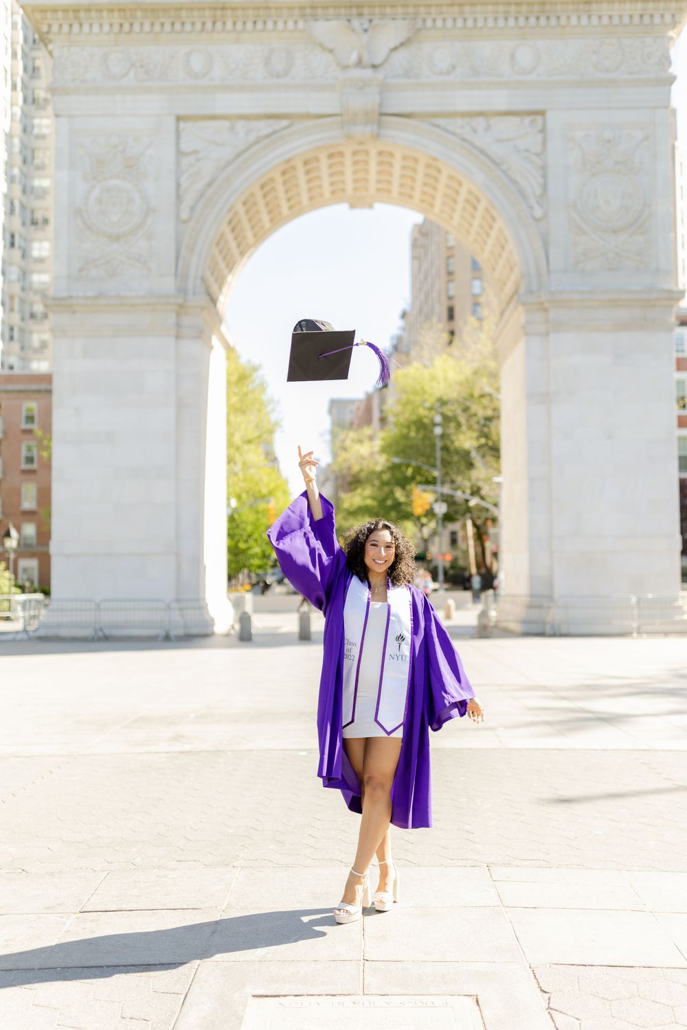 Graduate tosses cap in celebration in front of architectural arch on sunny day.