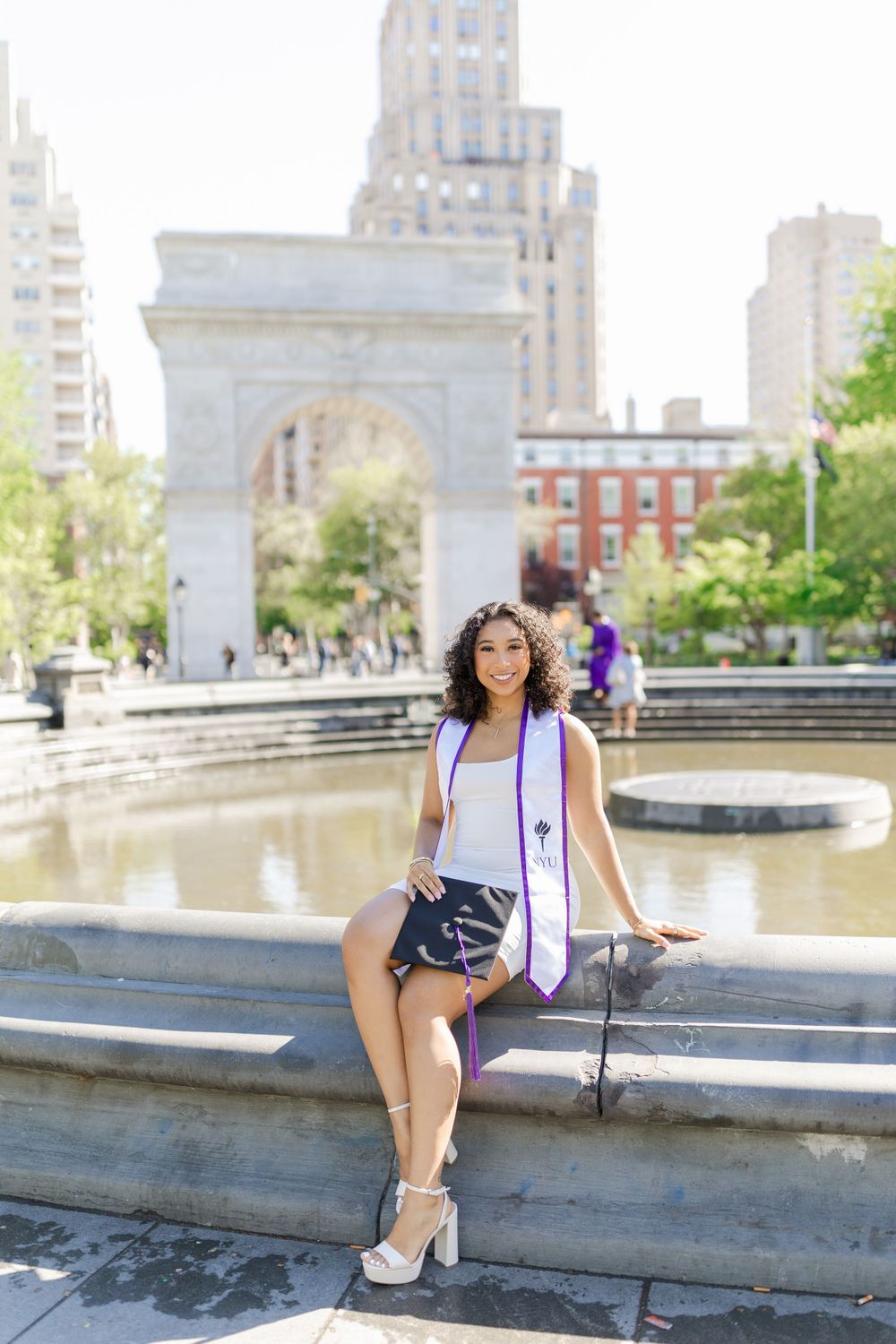 Graduate poses on stone ledge near fountain with Washington Square Arch in background.