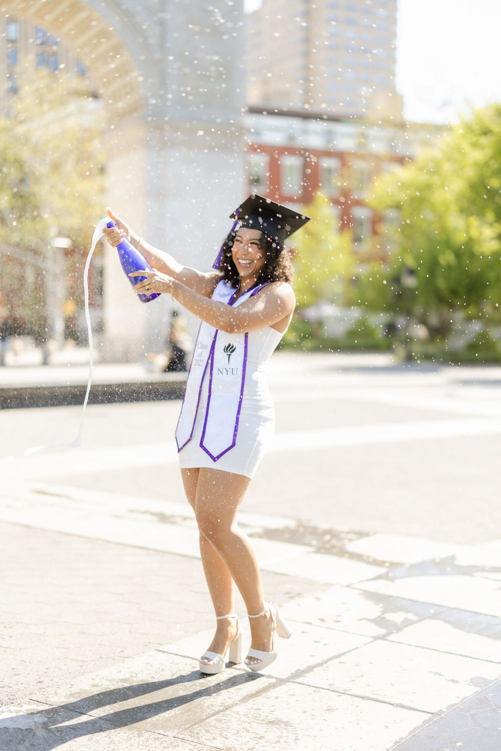 Graduate sprays champagne during celebration photoshoot outdoors.