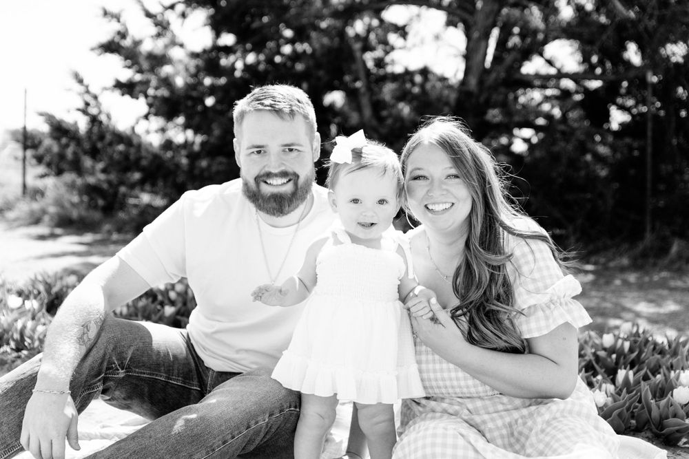 Black and white portrait of a family sitting outdoors together on a sunny day with trees in the background.