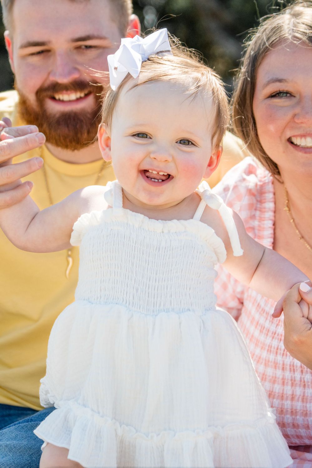 A joyful toddler in a white summer dress smiles brightly while being held by adults outdoors in sunlight.