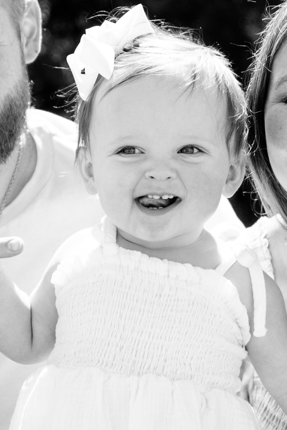 Happy toddler wearing white dress and bow smiles brightly in black and white portrait.