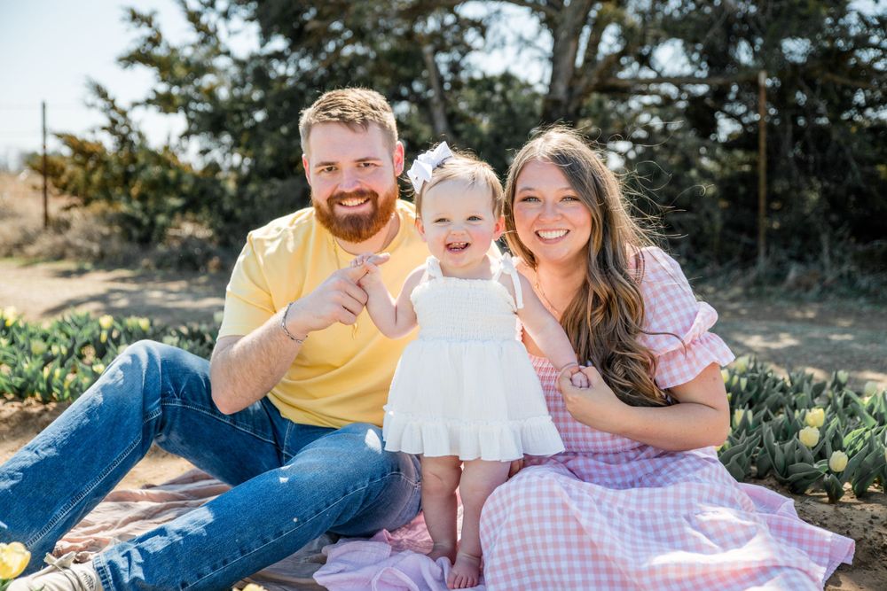 Happy family with toddler girl in white dress having outdoor photoshoot on pink blanket in sunny garden setting.