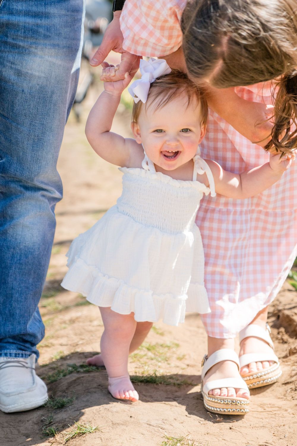 Baby wearing white dress smiles while walking barefoot and holding hands on a sunny day outdoors.