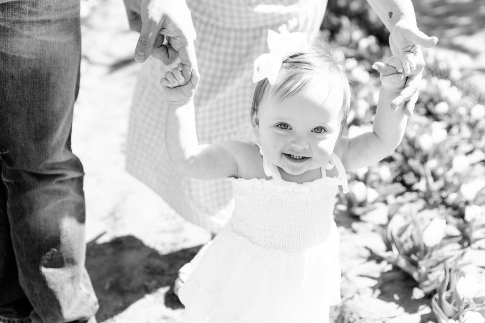 A child in a white dress smiles while holding hands with adults in a black and white outdoor photo.