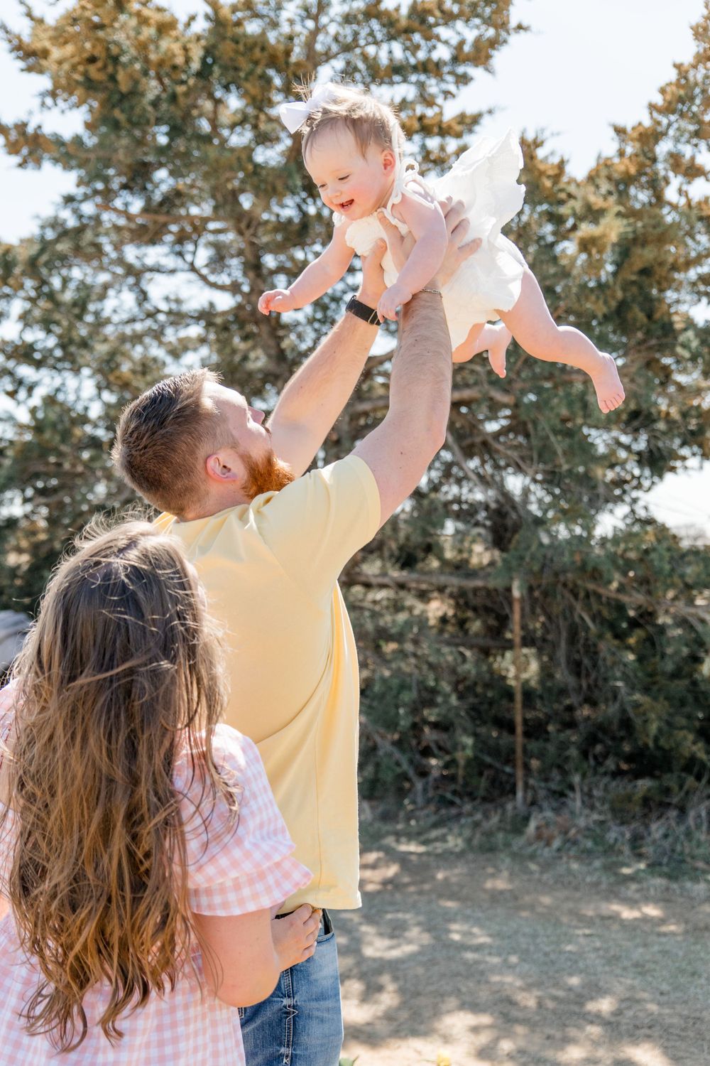 Joyful family moment outdoors as parent lifts laughing baby up high in the air on a sunny day near evergreen trees.