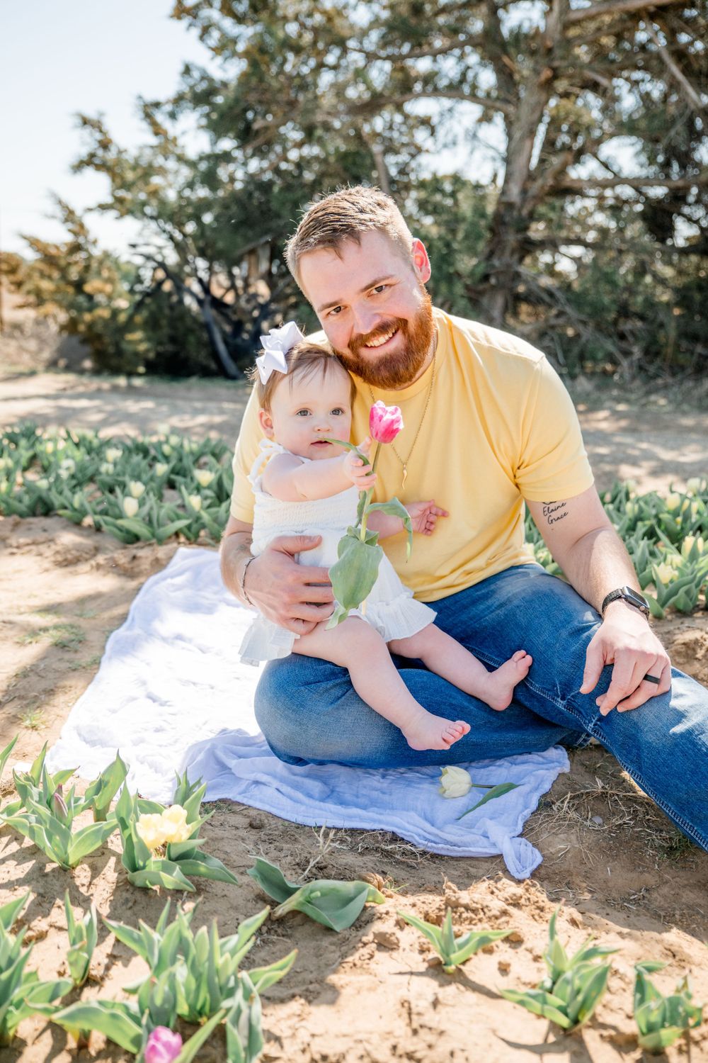 A parent and baby sit together on a blue blanket outdoors surrounded by greenery on a sunny day.