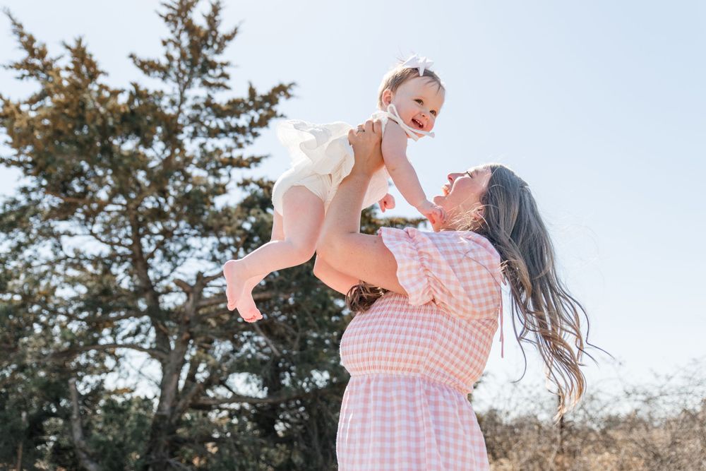 A parent lifts a joyful baby high in the air while wearing a pink gingham dress outdoors in front of evergreen trees.