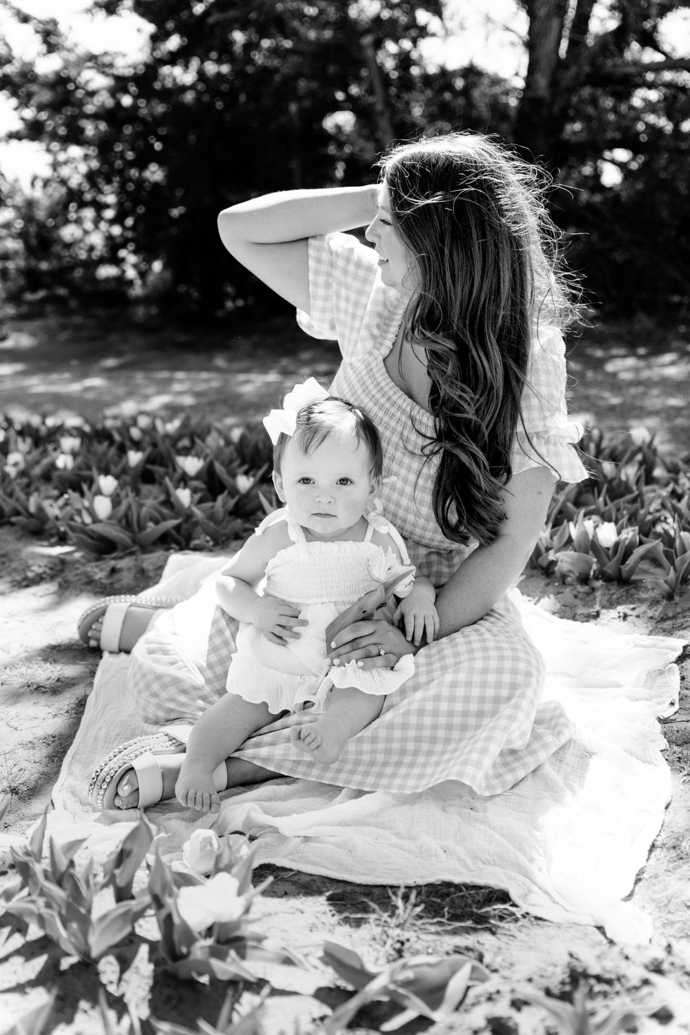 A black and white portrait of a parent and child sitting together on a blanket outdoors in dappled sunlight.