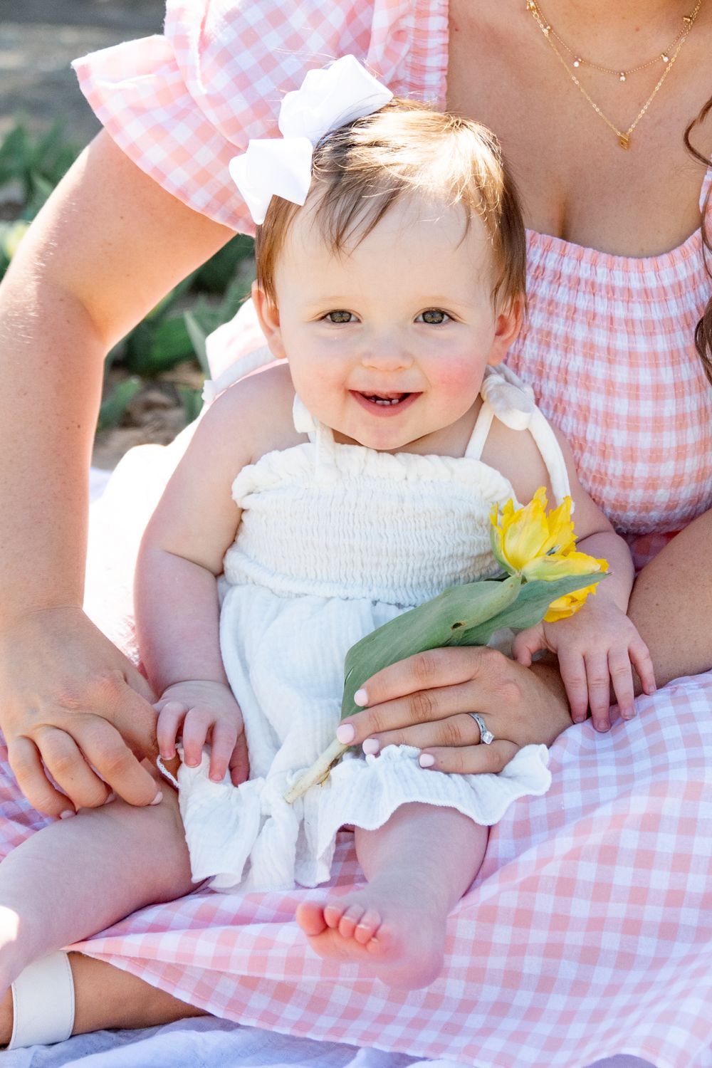 Sweet baby in white smocked dress holding yellow flower sits with parent wearing pink gingham dress on sunny day.