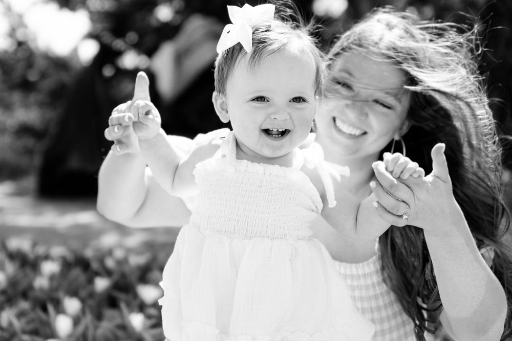 Black and white close-up of a baby in white dress and hair bow smiling with joy outdoors.