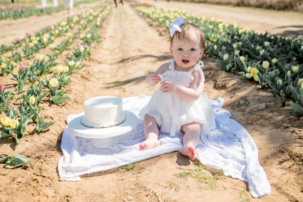 Small child in white dress sits on blanket surrounded by rows of tulip flowers in spring garden.