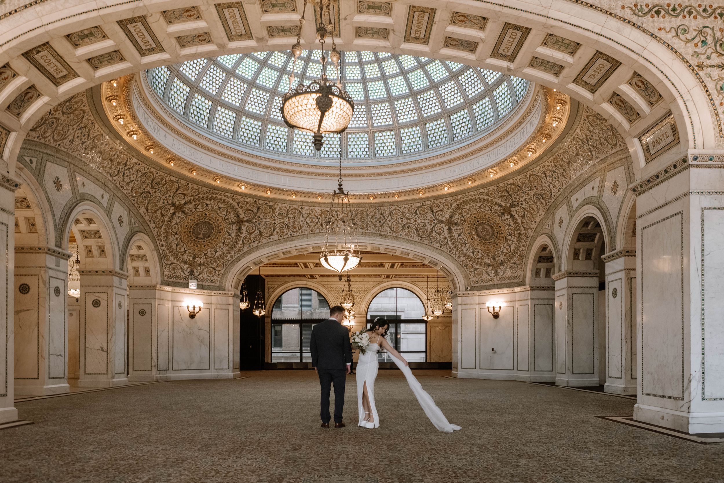 A couple in wedding attire stands beneath an ornate domed ceiling in the Chicago Cultural Center's historic Preston Bradley Hall.