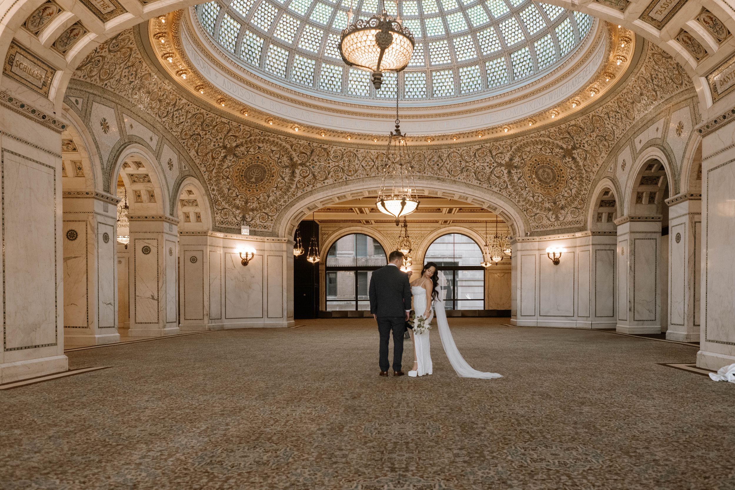 A couple in wedding attire stands beneath an ornate domed ceiling in a grand historic building with classical architecture.