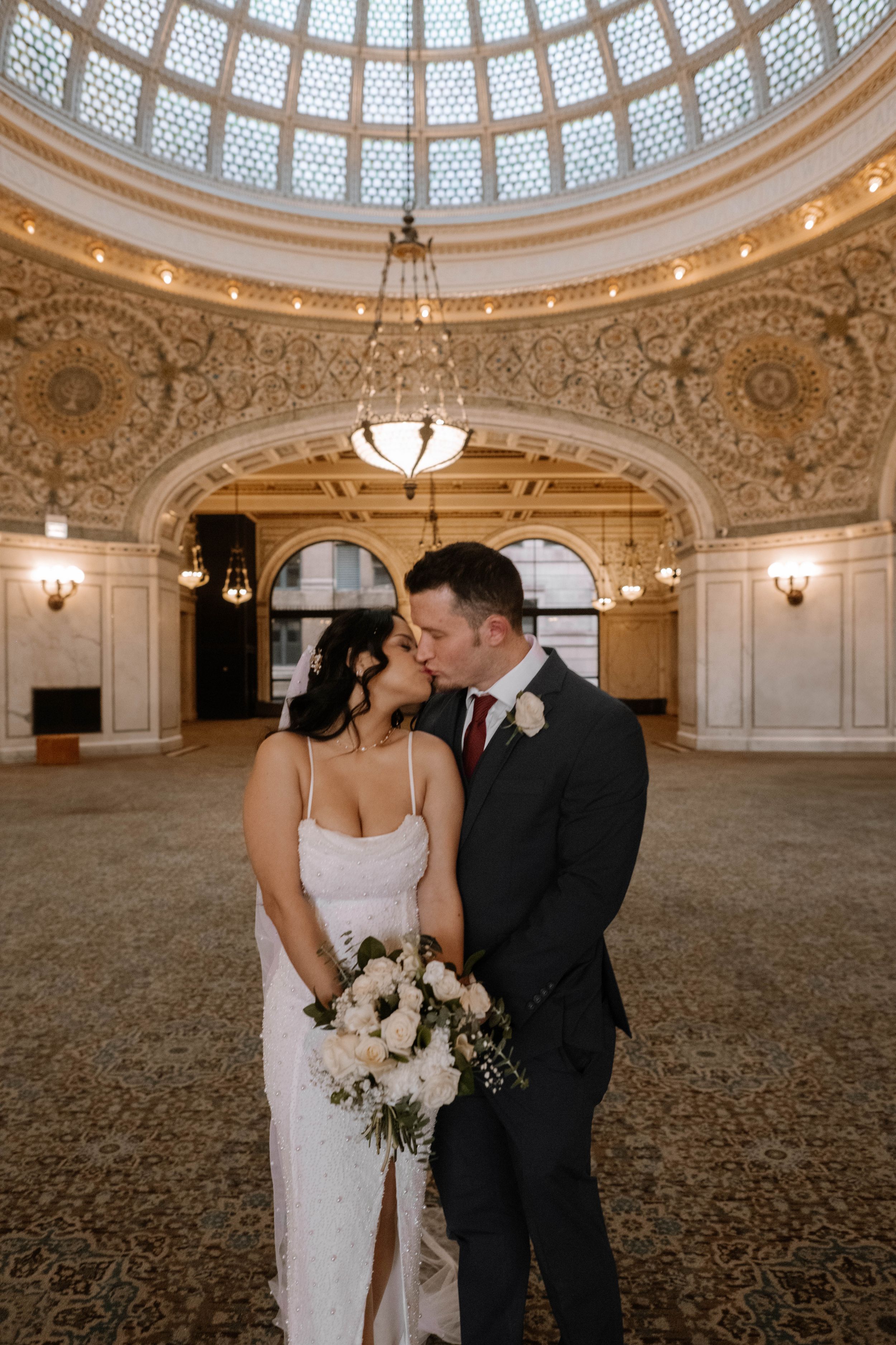 Elegant wedding couple shares romantic moment under ornate domed ceiling with chandelier at Chicago Cultural Center.