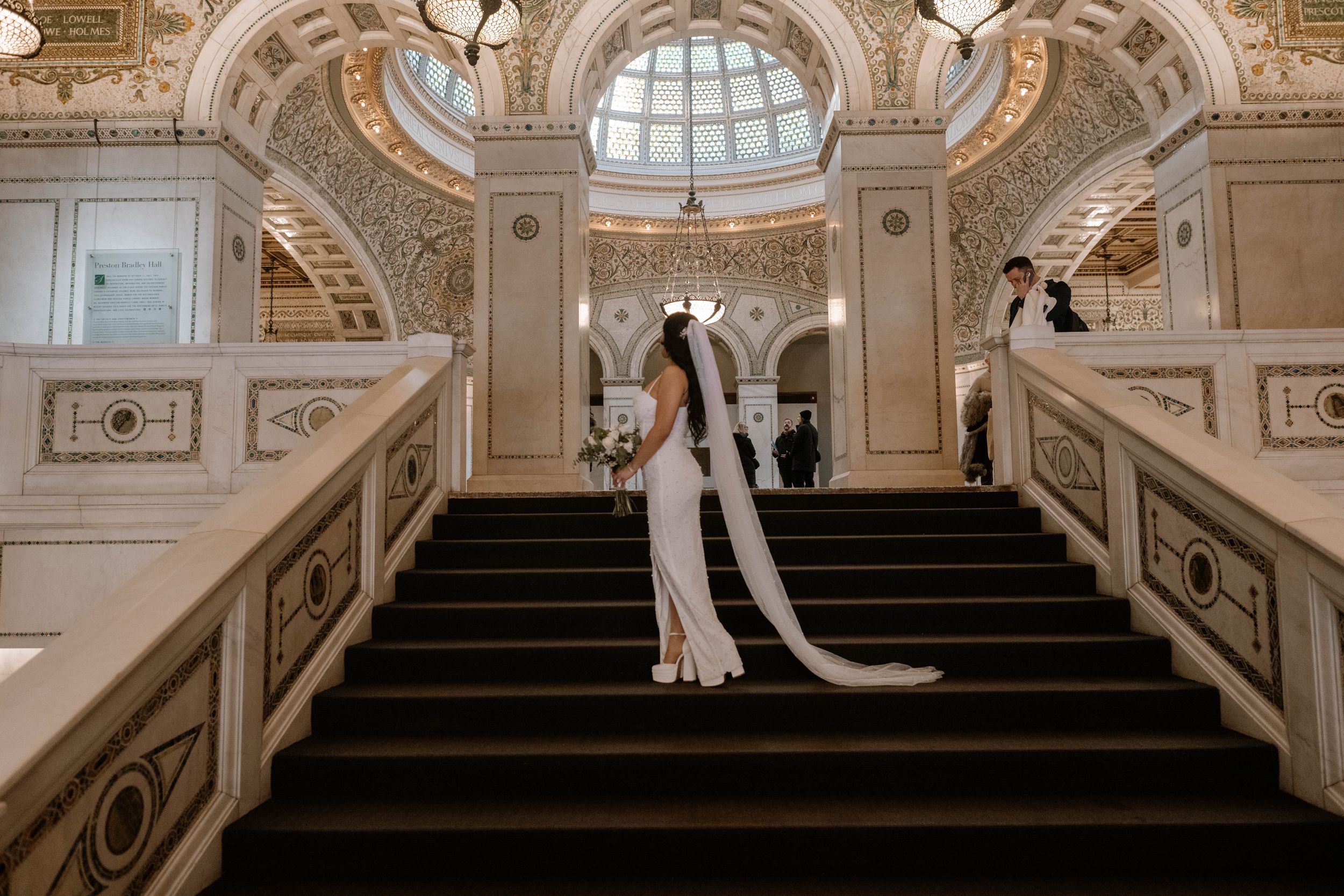 Elegant wedding scene on grand marble staircase inside ornate historic building with domed glass ceiling and baroque details.