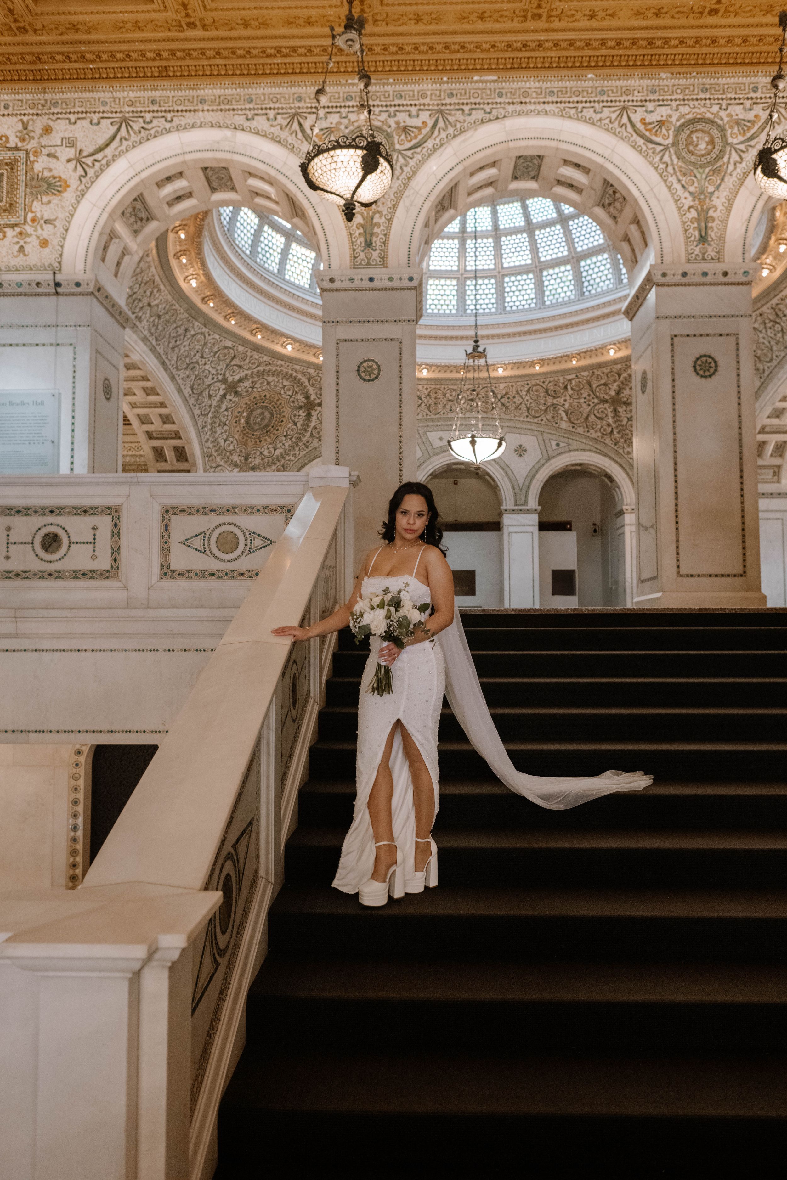 A bride in white wedding gown stands on grand staircase inside ornate historic building with domed ceiling and classic architecture.