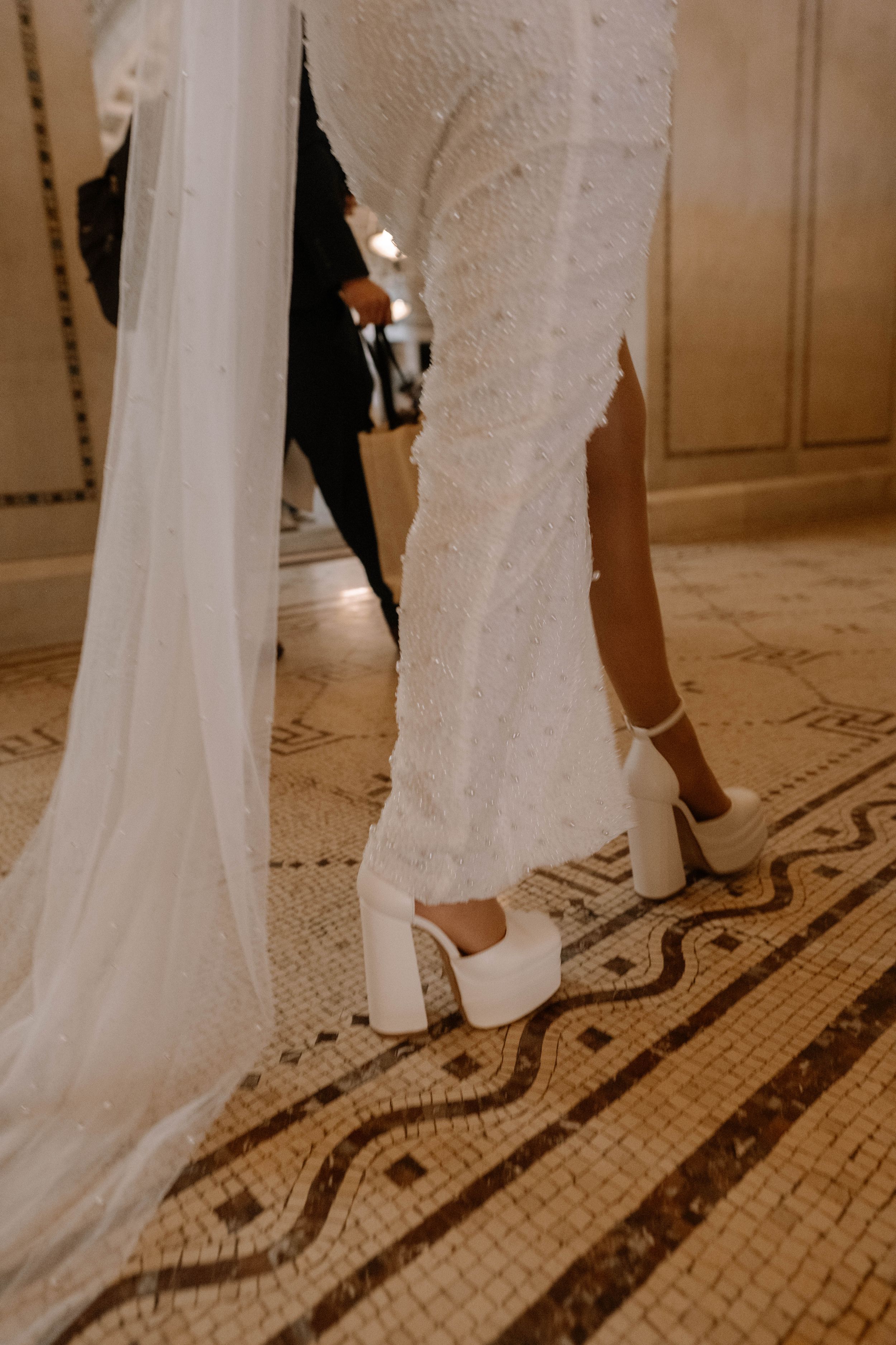 Close-up of bride wearing white platform heels and lace wedding dress hem on ornate mosaic tile floor.