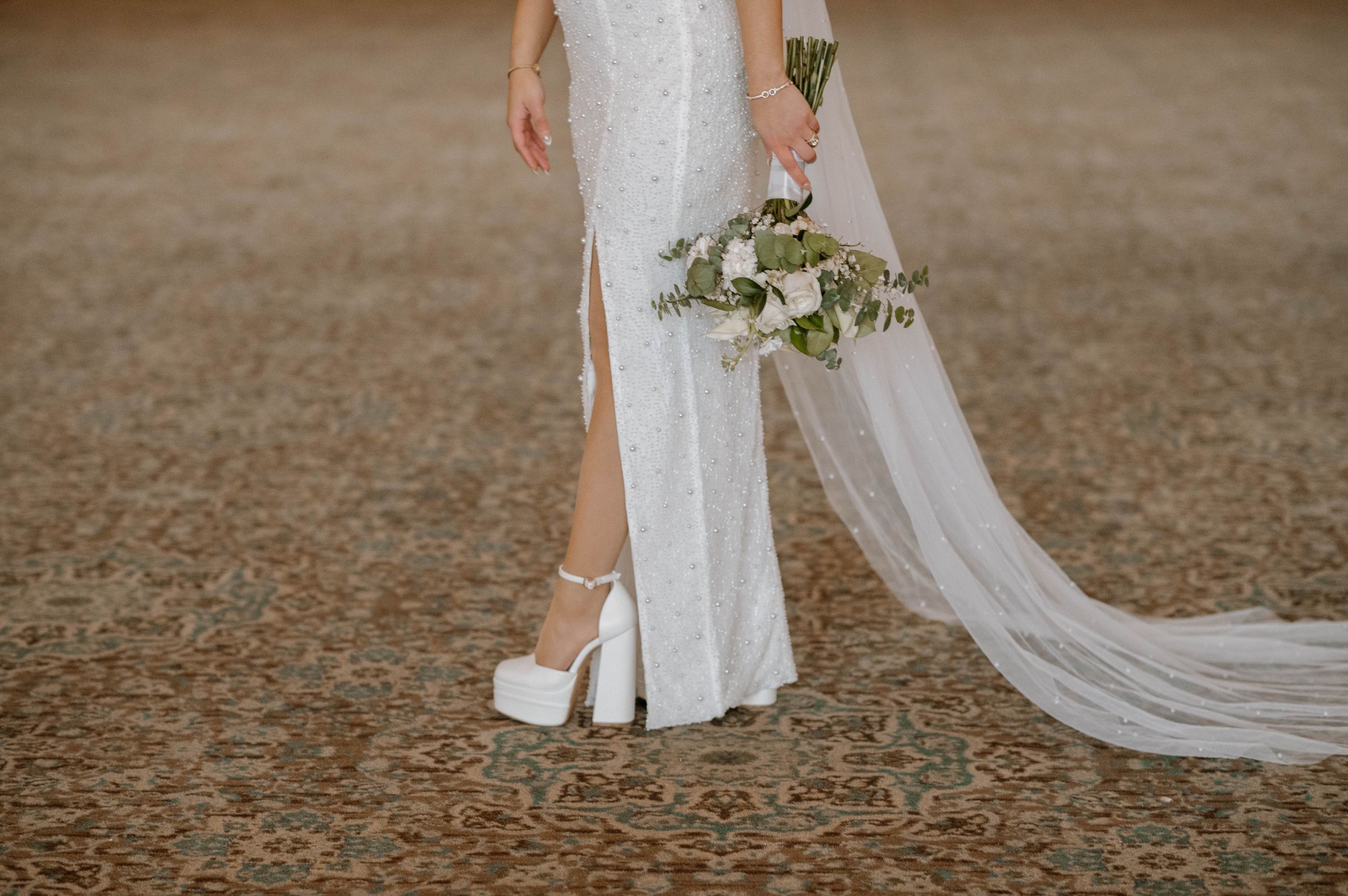 Close-up view of bride wearing white platform heels and lace dress with flowing veil holding floral bouquet.
