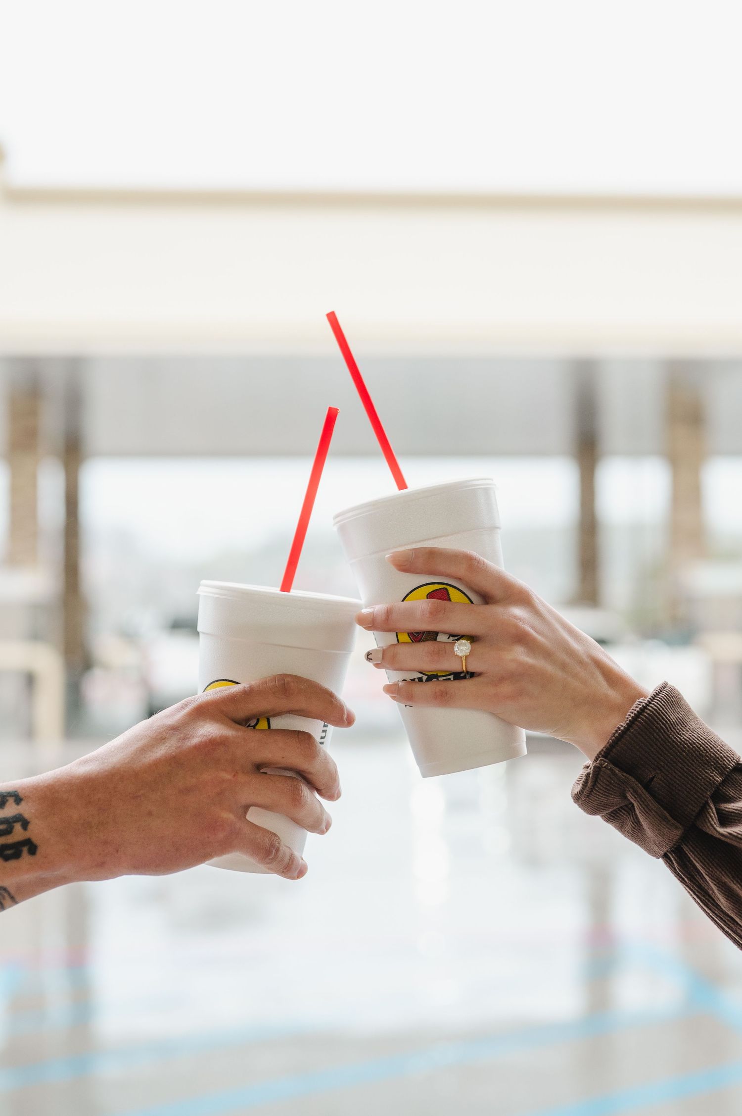 Two hands with tattoos toasting white disposable coffee cups with red straws against a bright background.
