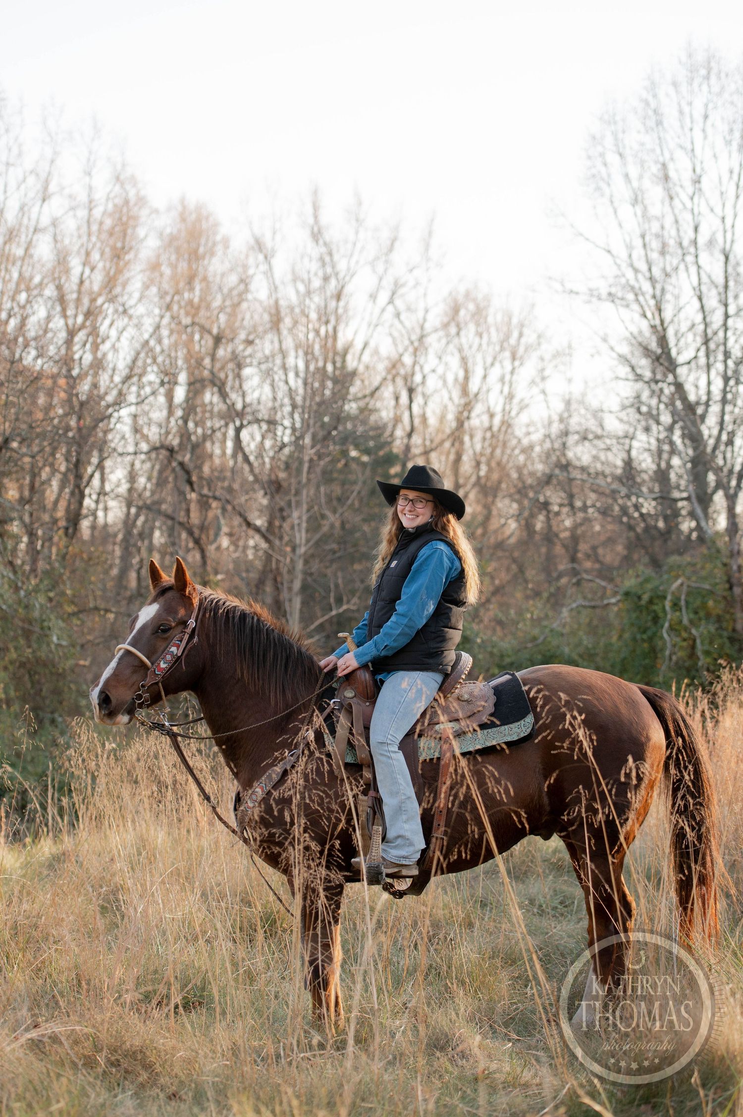Equestrian in blue jacket rides dark horse in winter field with bare trees.