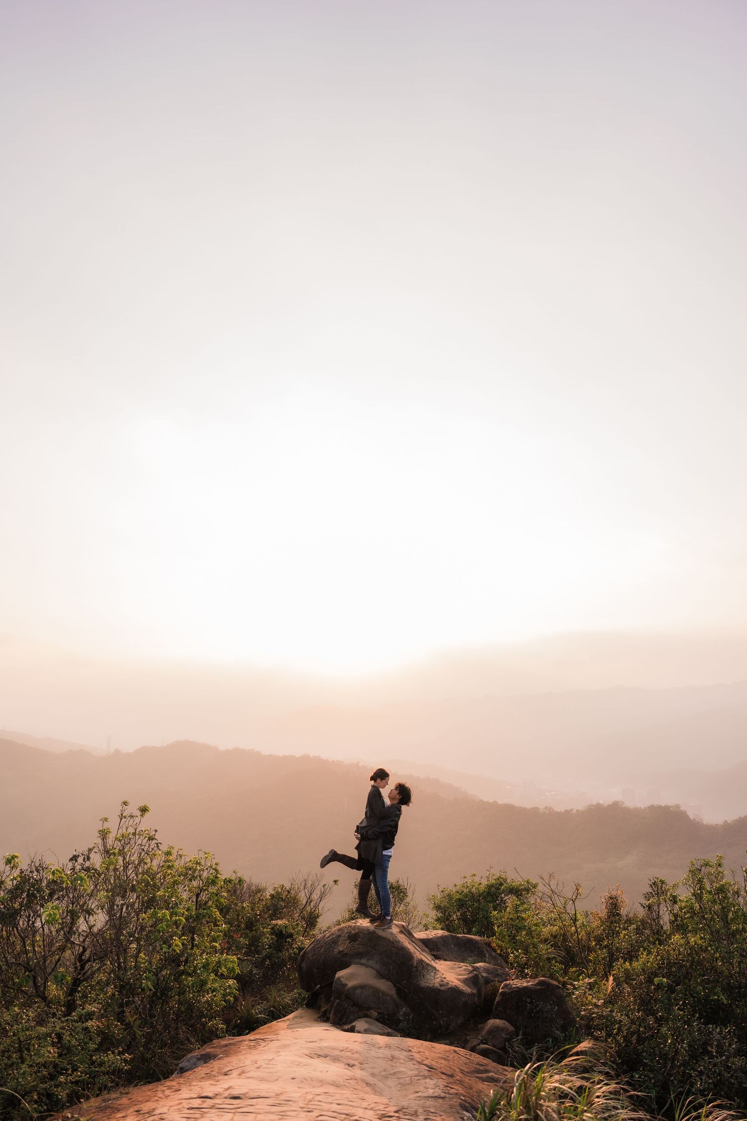 Two silhouettes stand together on a rocky overlook against a misty mountain landscape at sunset.