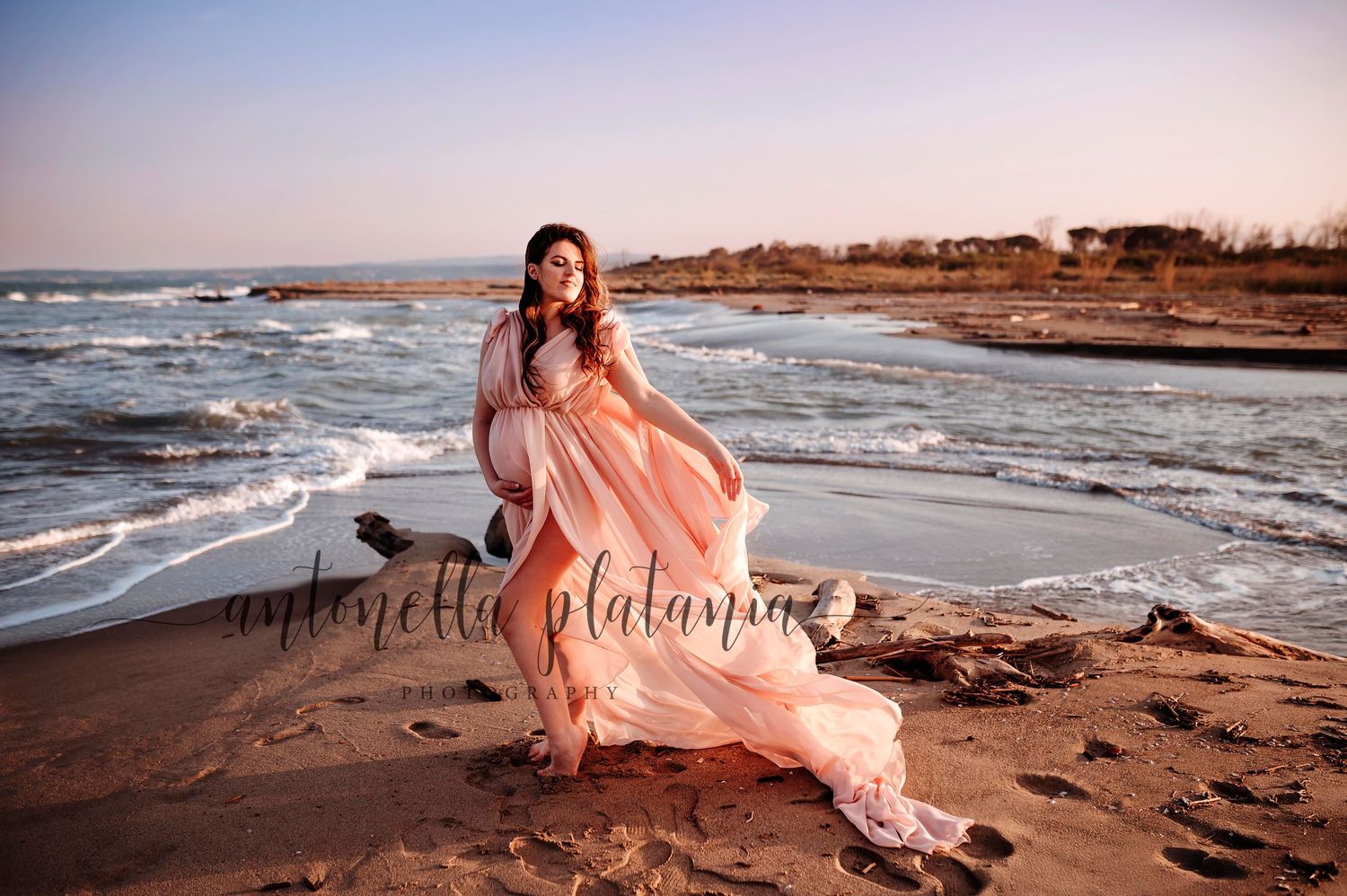 una donna incinta che posa con un vestito rosa in riva al mare, al tramonto. servizio fotografico maternità, catania
