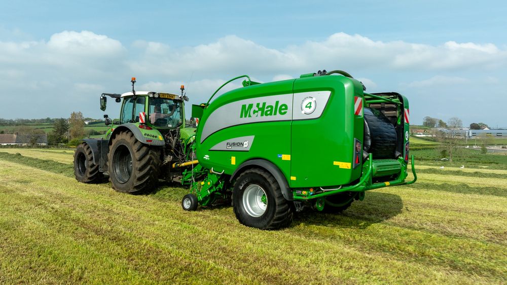 A green McHale baler machine and tractor working together in a grassy agricultural field on a sunny day.