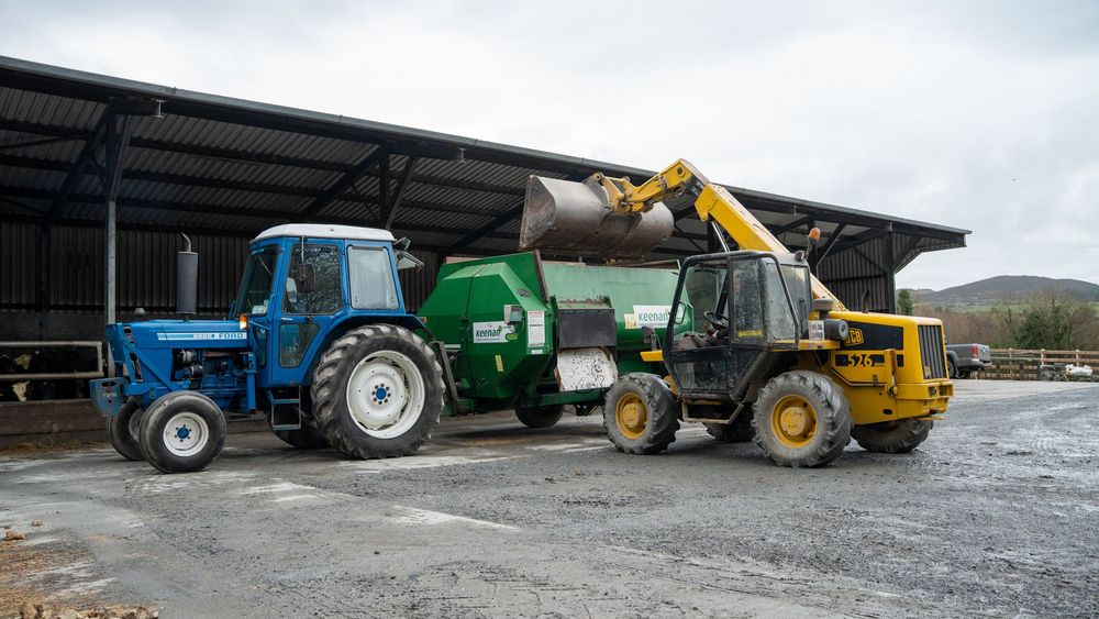 A panoramic view of farm equipment and tractors parked in a gravel lot with mountains in the background.