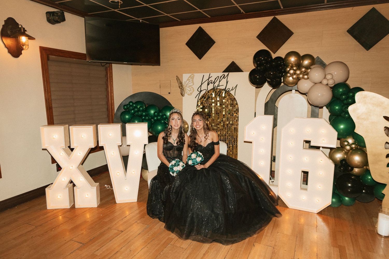 Elegant celebration setup with large white XV letters, black tulle dress, and green and gold balloon arch decorations.