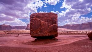 Massive red rock boulder sits in desert landscape under dramatic cloudy sky.