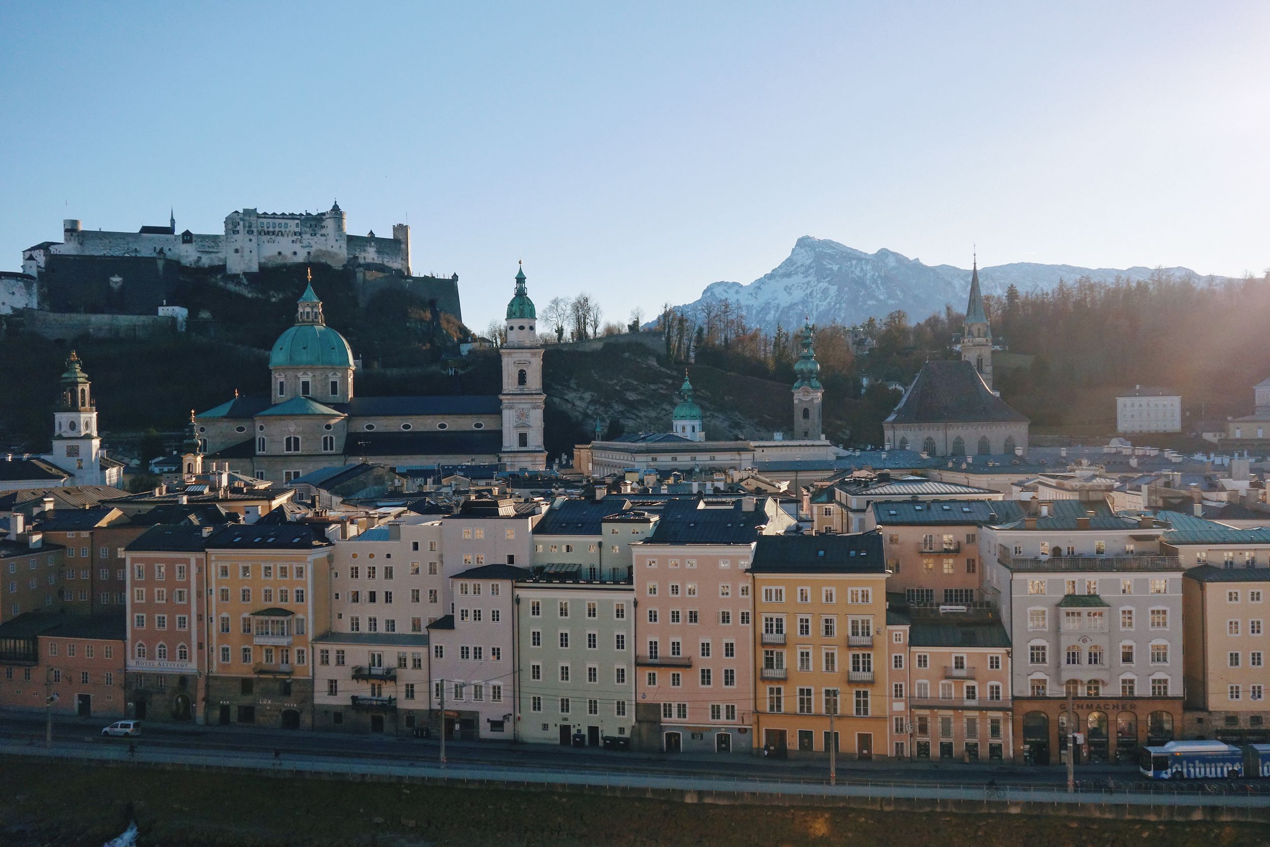 Historic cityscape of Salzburg, Austria with Hohensalzburg Fortress on hilltop and pastel buildings along riverbank at sunset.