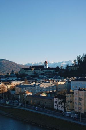 Historic cityscape of Salzburg with church spires and mountains in background along river at sunset.