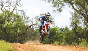 Motocross rider catching air during jump on a dirt track with trees in background.